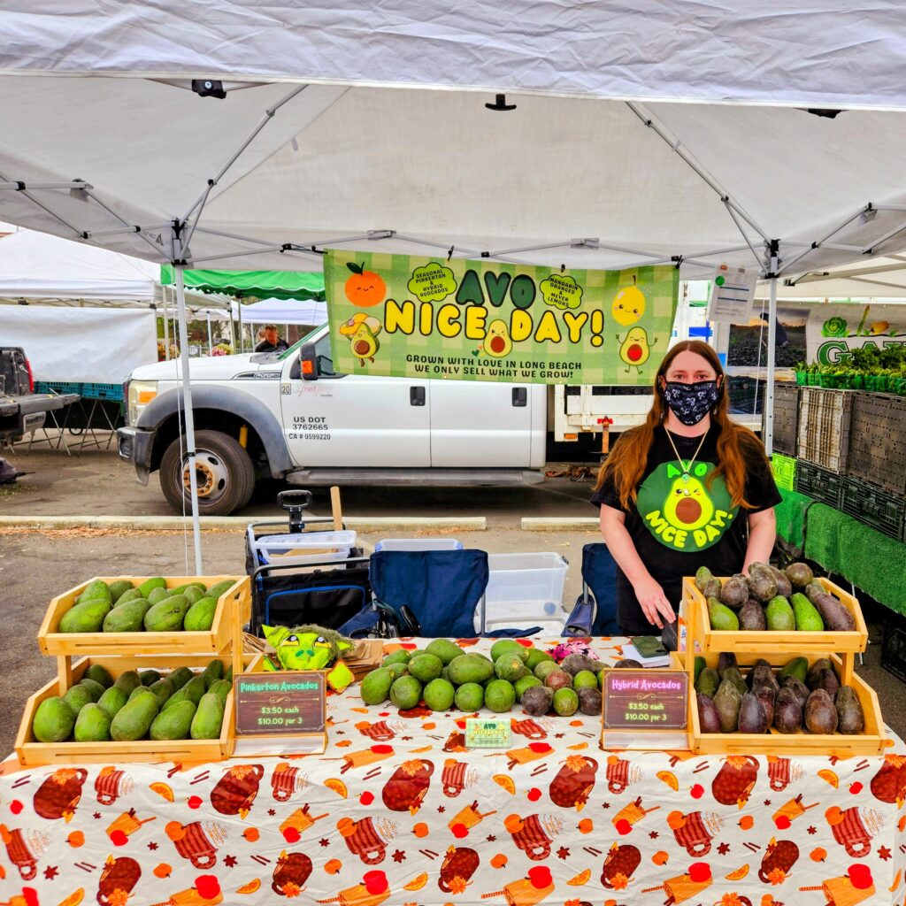 Market stall under a white tent selling crates of avocados with a colorful 'Avo Nice Day' sign and vendor wearing a mask.