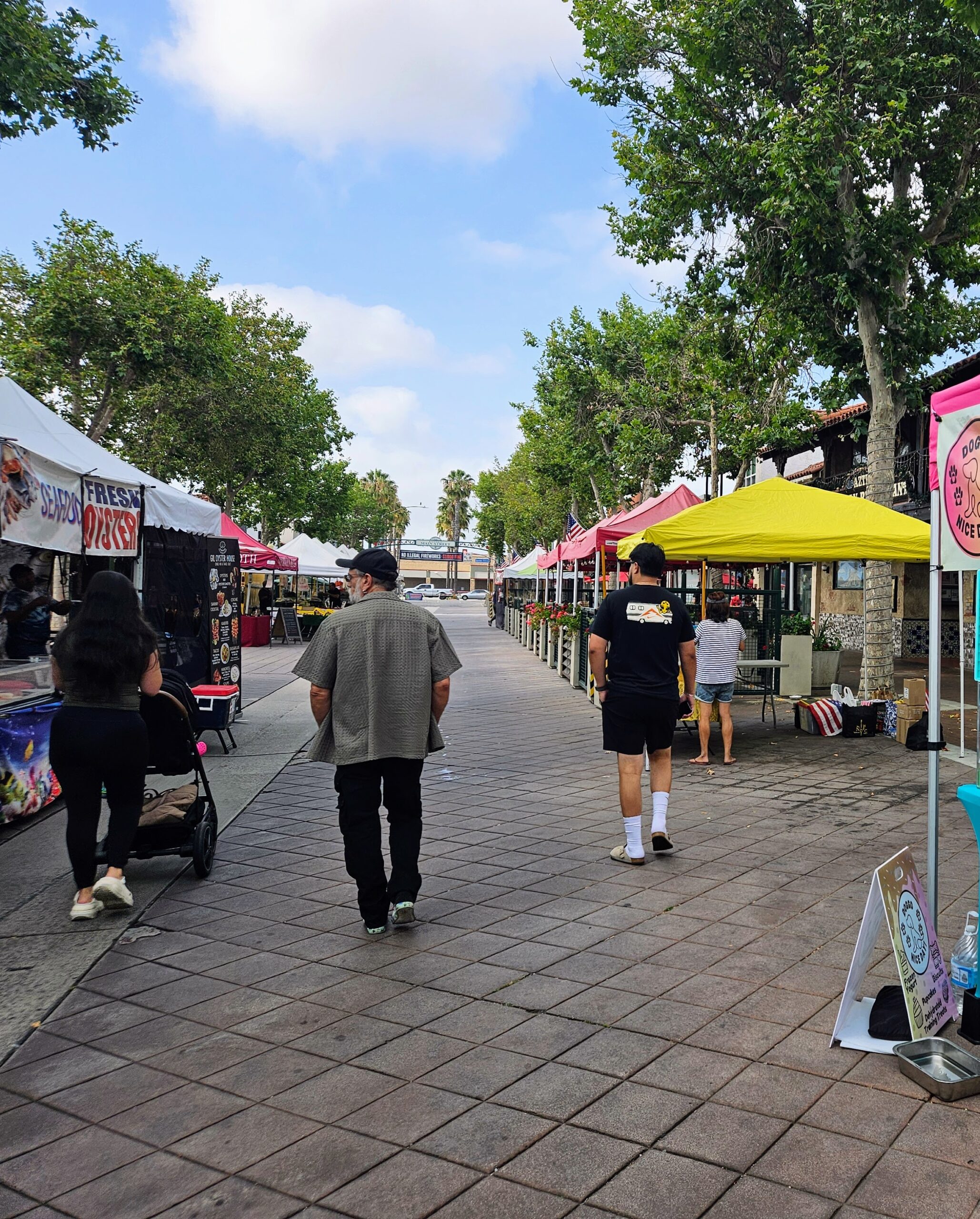 Street view of an outdoor farmers market with colorful vendor tents and people walking down the aisle.