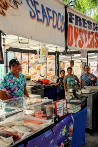 Farmers market seafood stall with fresh fish and oysters on display and smiling vendors under a canopy.