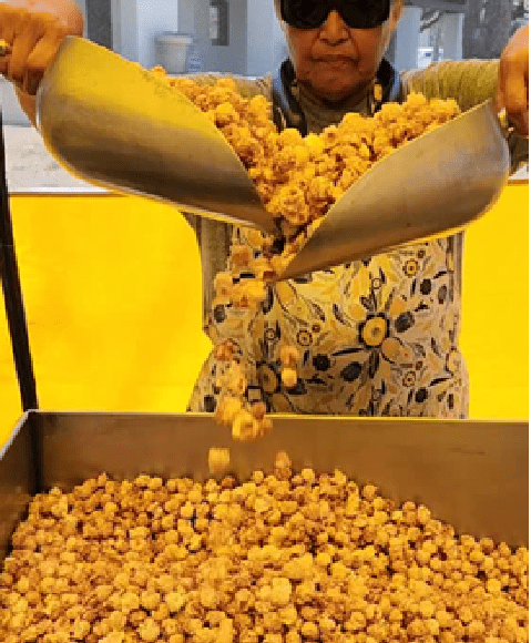 Vendor scooping caramel popcorn from metal scoops into large tray at a market stall.