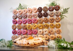 Assorted glazed and decorated donuts arranged on a vertical donut wall with a wooden platter of pastries in front and greenery accents.
