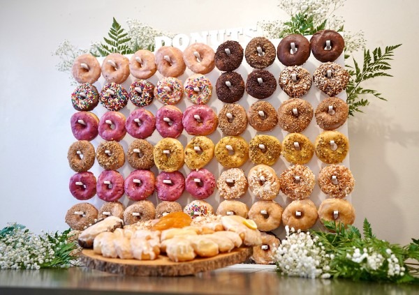 Assorted glazed and decorated donuts arranged on a vertical donut wall with a wooden platter of pastries in front and greenery accents.