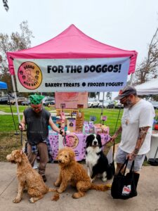 Outdoor market booth with a pink tent banner reading 'For the Doggos!' and three dogs with two handlers.