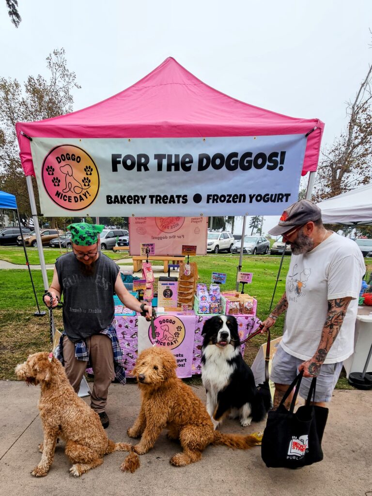 Outdoor market booth with a pink tent banner reading 'For the Doggos!' and three dogs with two handlers.