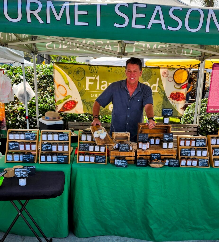 Vendor standing behind a farmers market stall displaying jars of spices and small chalkboard labels beneath a green canopy.