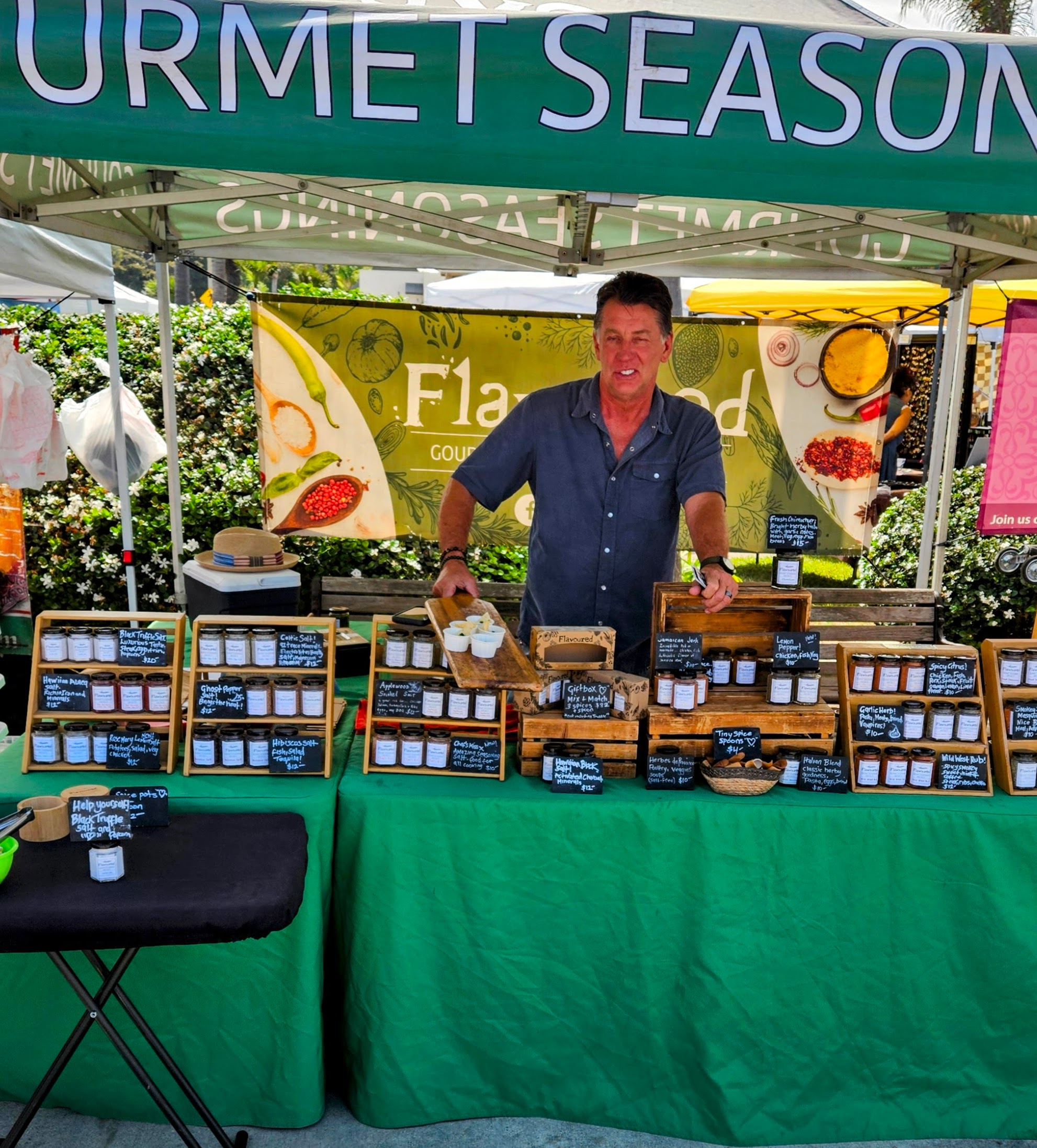 Vendor standing behind a farmers market stall displaying jars of spices and small chalkboard labels beneath a green canopy.