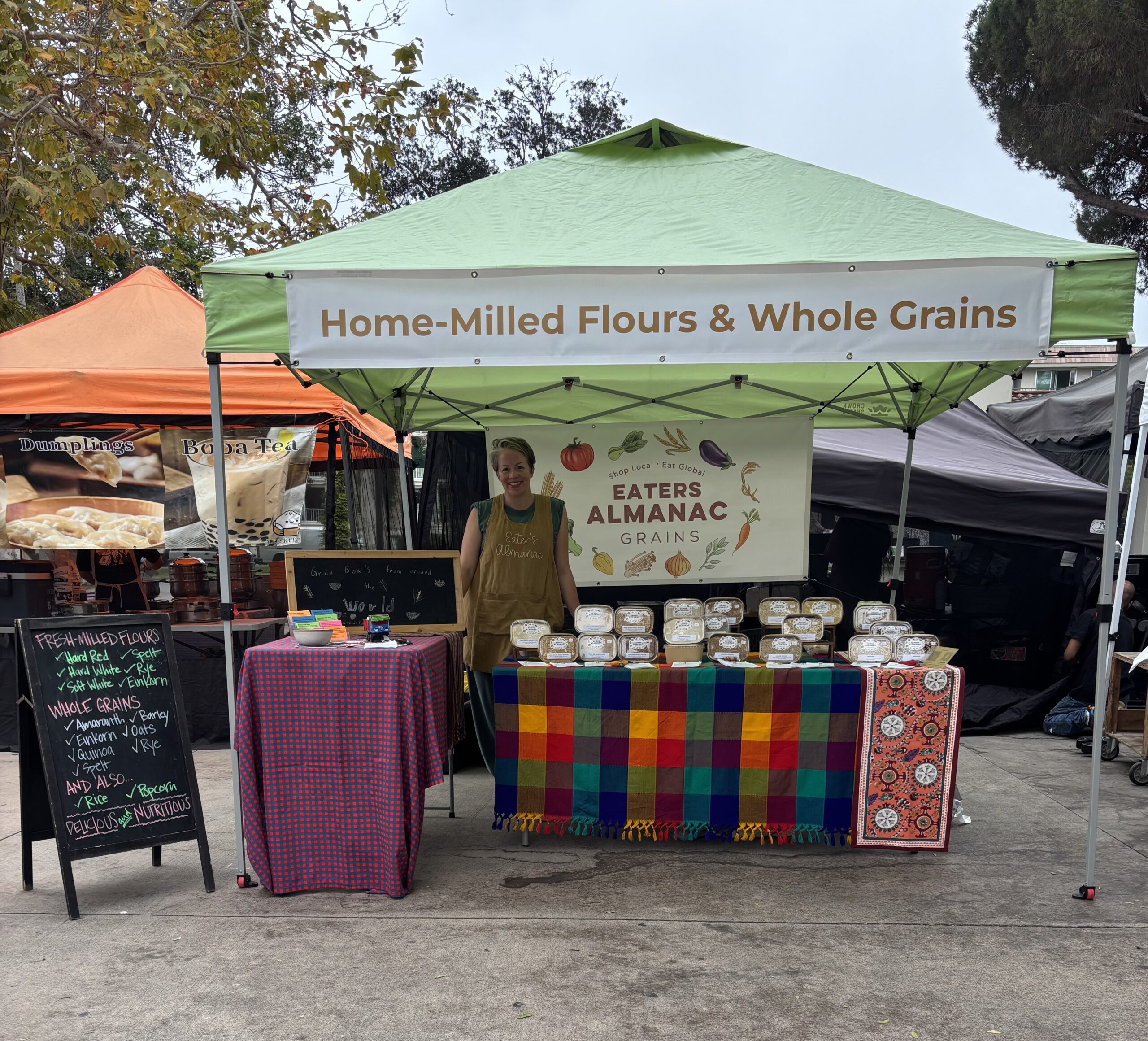 Vendor stall under a green canopy with packaged grains and a chalkboard sign at a farmers market.