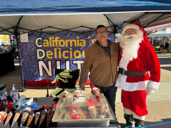 Vendor and person dressed as Santa Claus standing at a nuts booth at an outdoor farmers market tent.