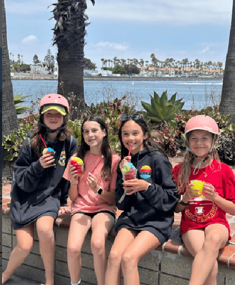 Four girls sitting on a low wall by the water eating colorful shaved ice treats with palm trees in the background.