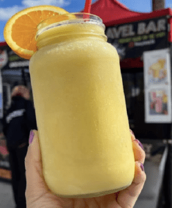 Hand holding a yellow smoothie in a mason jar topped with an orange slice and red straw at an outdoor market stall.