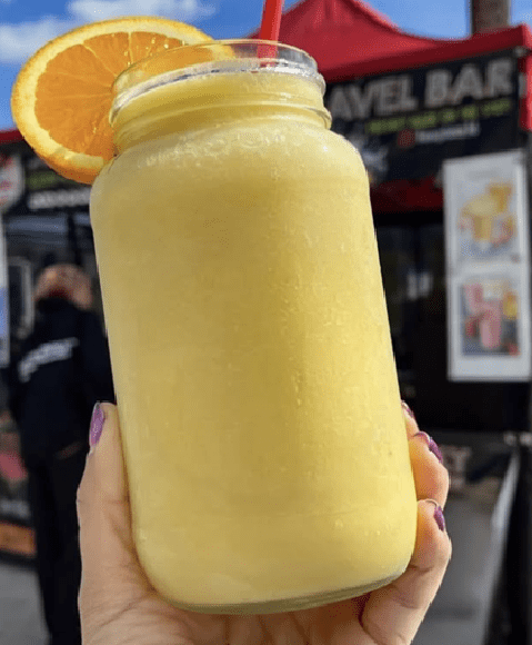 Hand holding a yellow smoothie in a mason jar topped with an orange slice and red straw at an outdoor market stall.