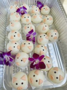 Tray of white steamed buns decorated like pig faces with small orange features and purple orchid flowers.