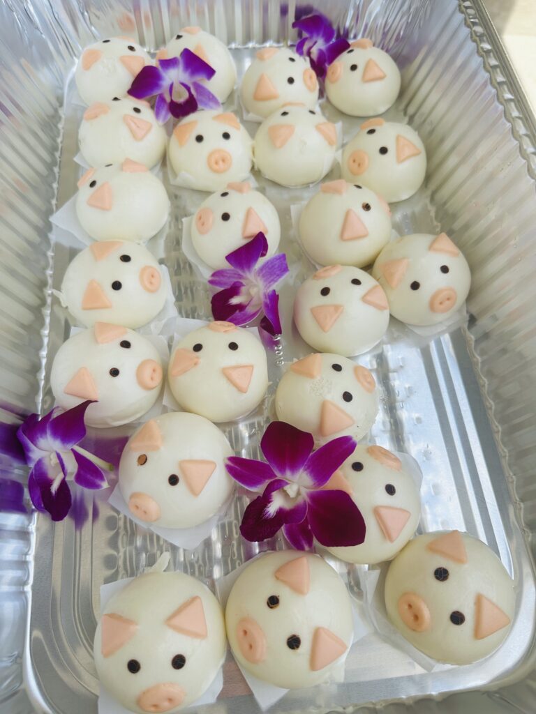 Tray of white steamed buns decorated like pig faces with small orange features and purple orchid flowers.