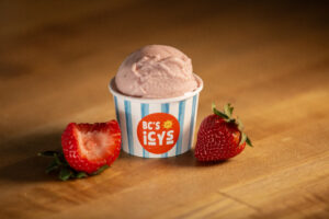 Single scoop of pink strawberry ice cream in a striped paper cup with fresh strawberries on a wooden board.