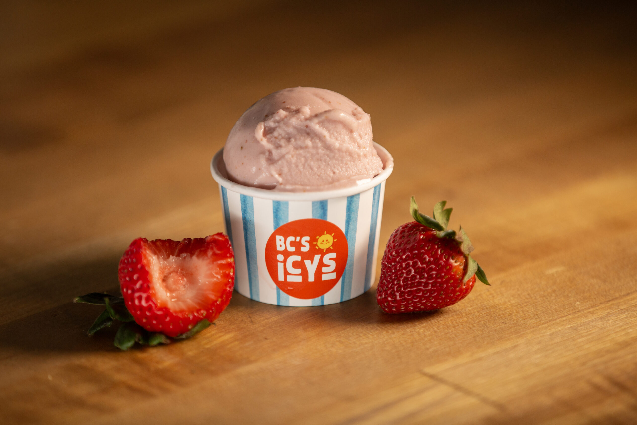 Single scoop of pink strawberry ice cream in a striped paper cup with fresh strawberries on a wooden board.
